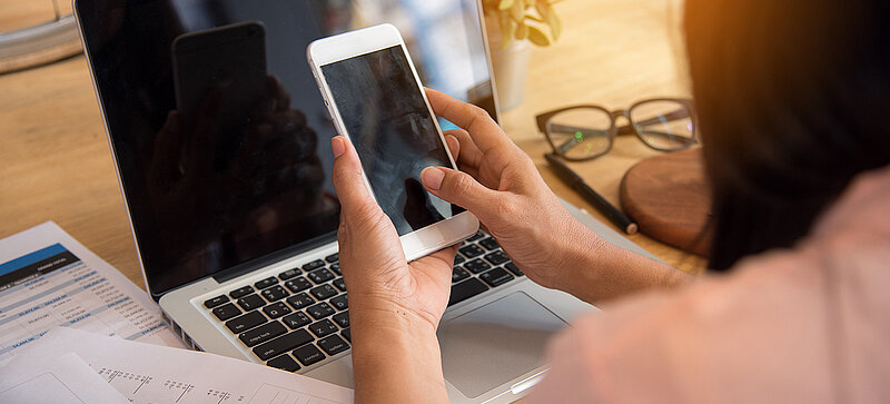 Eine Frau sitzt an einem Schreibtisch mit einem Laptop und hält ein Smartphone in der Hand